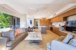 A modern living room with a sofa, coffee table, and a TV. There is a kitchen area with wooden cabinets and a large TV.