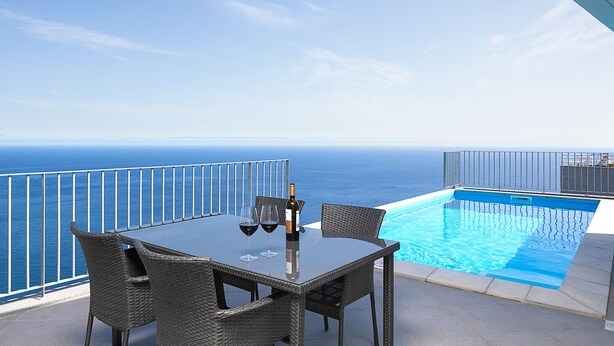 Une table à plateau en verre, des chaises en osier, une bouteille de vin et deux verres à vin, le tout sur un balcon donnant sur une piscine et l’océan.
