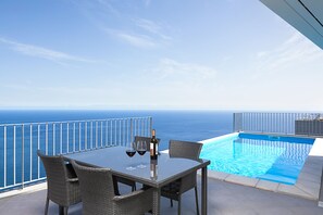 A glass-top table with wicker chairs, a bottle of wine, and two wine glasses on a balcony overlooking a pool and the ocean.