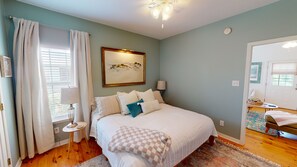 Un dormitorio con cama, una mesita de noche con lámpara, una ventana con cortinas, un cuadro en la pared y una puerta que da a otra habitación.
