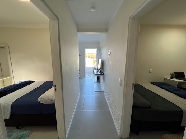 A corridor leading to a bedroom with two beds, a desk with a laptop, and a small plant.