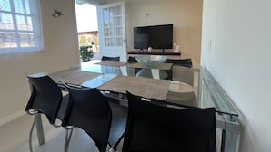Comedor con mesa de vidrio, sillas negras, televisor y puerta que da al exterior.