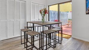 Une pièce avec une table en bois et quatre tabourets, une porte-fenêtre coulissante donnant sur un espace extérieur délimité par une clôture en bois et sur lequel se trouve un objet rouge.