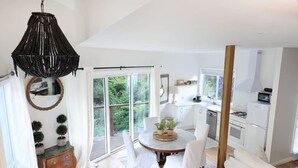 A modern kitchen with white cabinets, a dining area with a round table and chairs, a hanging black chandelier, and a sliding glass door leading to a deck. - Sunny Corner - Romantic Split Level Cottage (Leura)