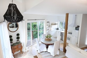 Une cuisine moderne avec des meubles blancs, un coin repas avec une table ronde et des chaises, un lustre noir suspendu et une porte-fenêtre coulissante donnant sur une terrasse.