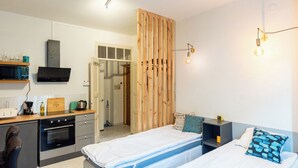A compact hotel room with a kitchenette, a bed with a blue and white bedspread, a wooden partition, and a wall-mounted TV. - HAPPY HOME HELSINKI-KALLIO (Helsinki)