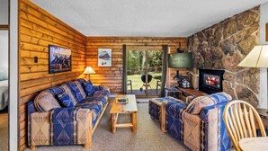 Interior acogedor de una cabaña con pared de piedra, mobiliario de madera, chimenea y vistas al exterior.