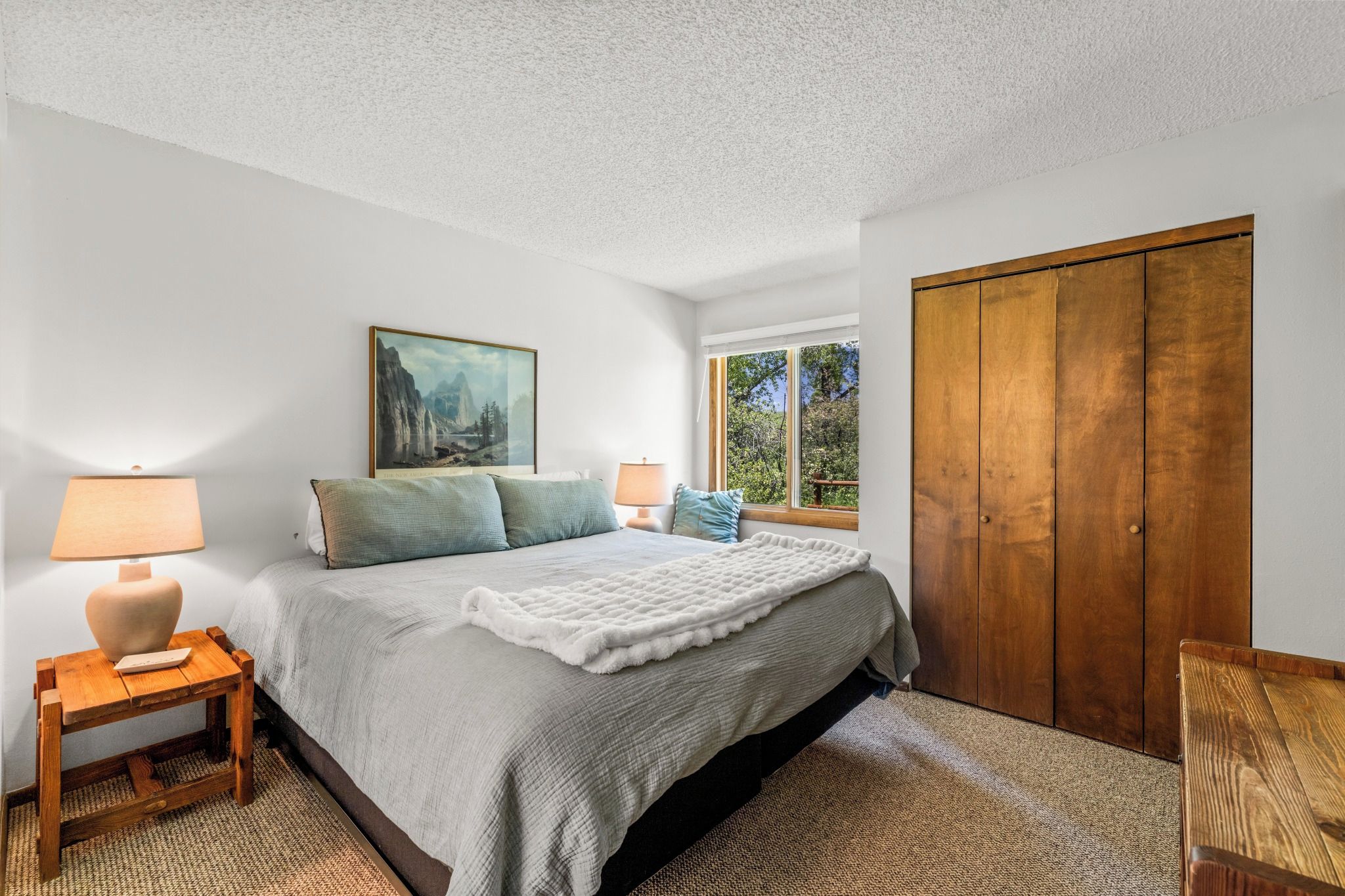 A bedroom with a bed, a nightstand, a lamp, a painting, a window, and a wardrobe.