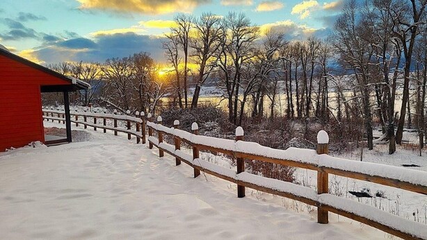 Una valla de madera cubierta de nieve que conduce a un edificio rojo, con árboles desnudos y una puesta de sol al fondo.