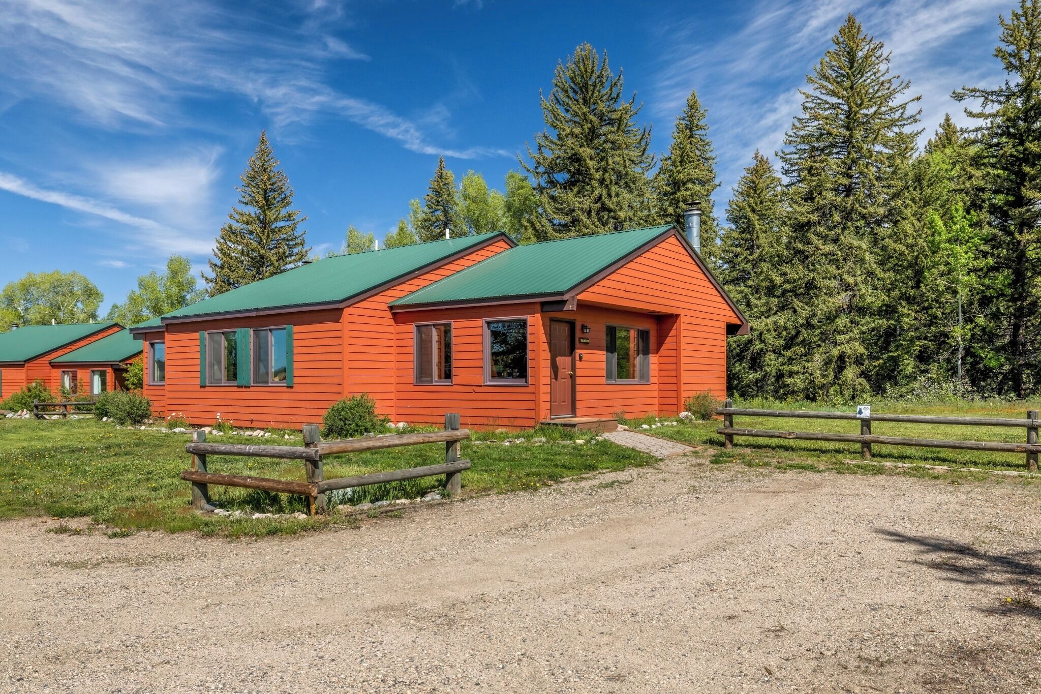 A red cabin with a green roof, surrounded by a gravel path and a wooden fence, set in a forested area.