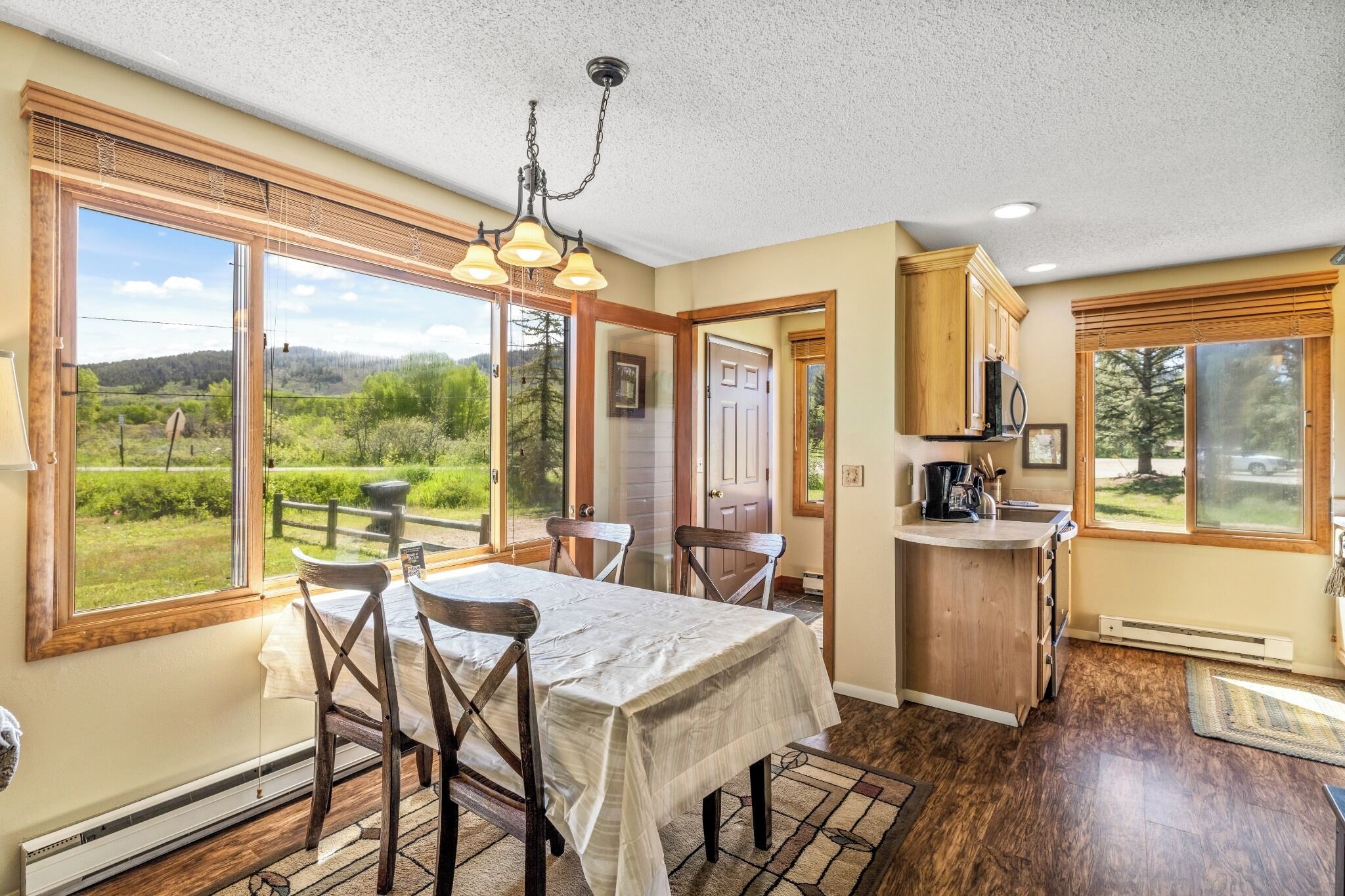 A dining area with a table and chairs, a kitchen with wooden cabinets, and large windows offering a view of the outdoors.