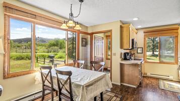 A dining area with a table and chairs, a kitchen with wooden cabinets, and large windows offering a view of the outdoors.