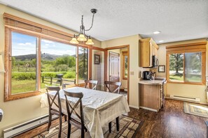 A dining area with a table and chairs, a kitchen with wooden cabinets, and large windows offering a view of the outdoors. - Pioneer - Cozy riverside retreat in the mountains (Clark)