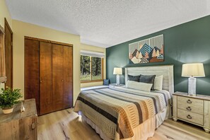 A bedroom with a large bed, two bedside tables with lamps, a wooden wardrobe, and a window with a view of trees.