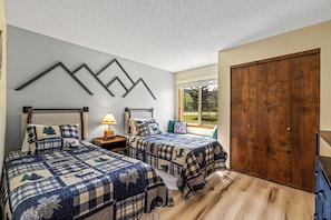 A bedroom with two beds, a wooden wardrobe, and a window with a view of trees.