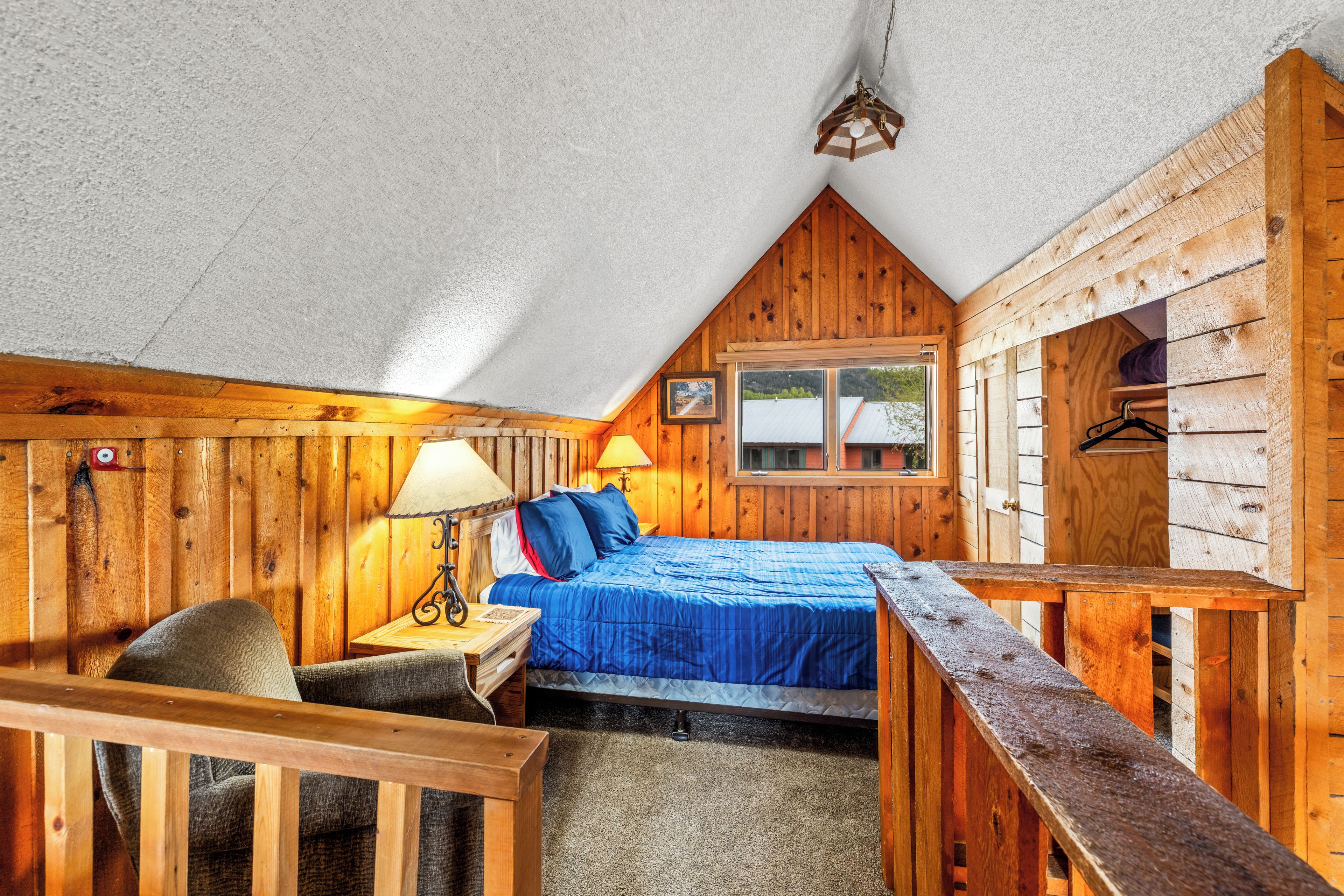 A cozy attic bedroom with a sloped ceiling, wooden walls, a bed with blue bedding, a desk with a lamp, and a window with a view of the outside.