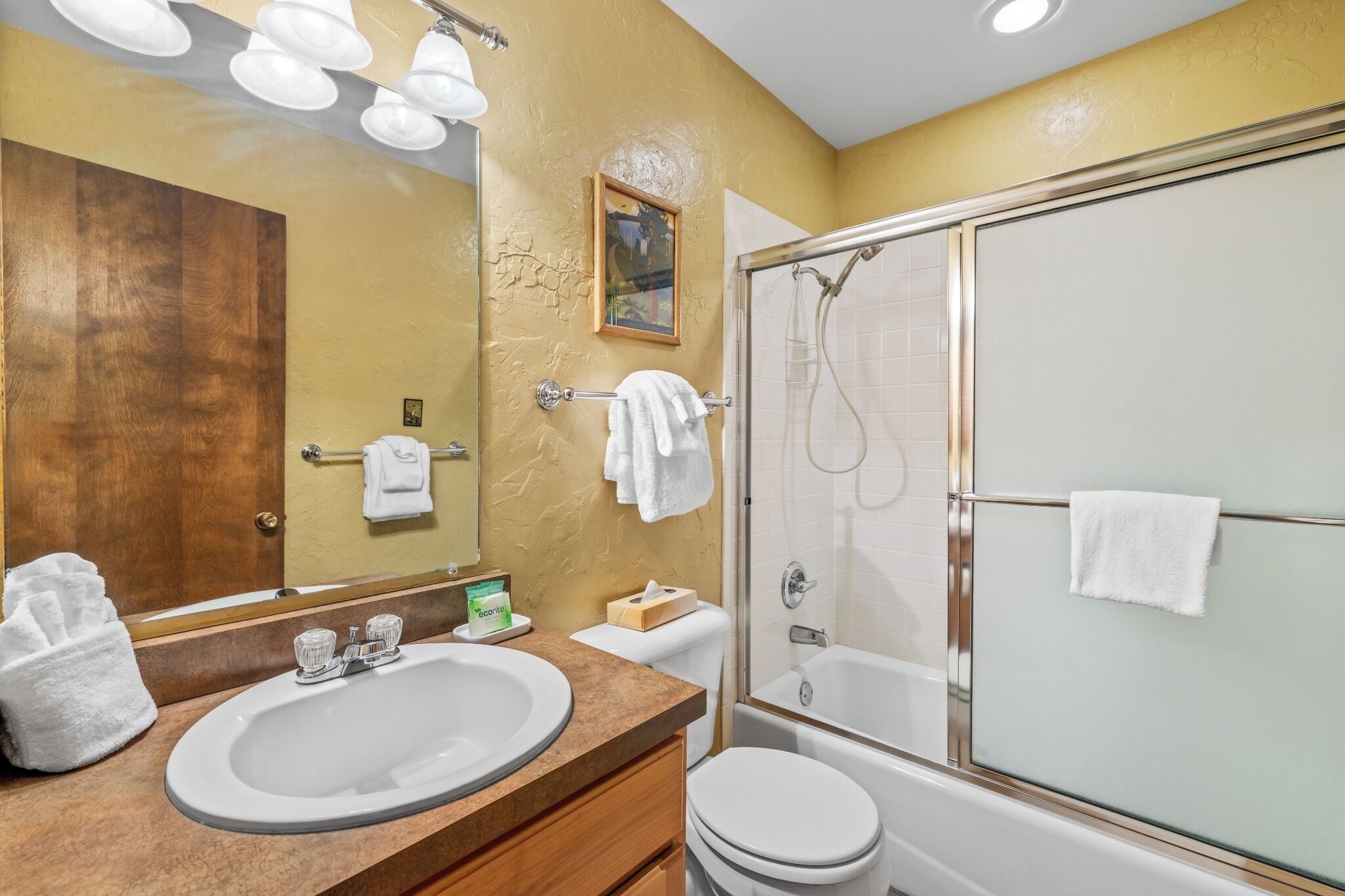 A bathroom with a double sink, a toilet, a glass-enclosed shower, and a mirror with lights.