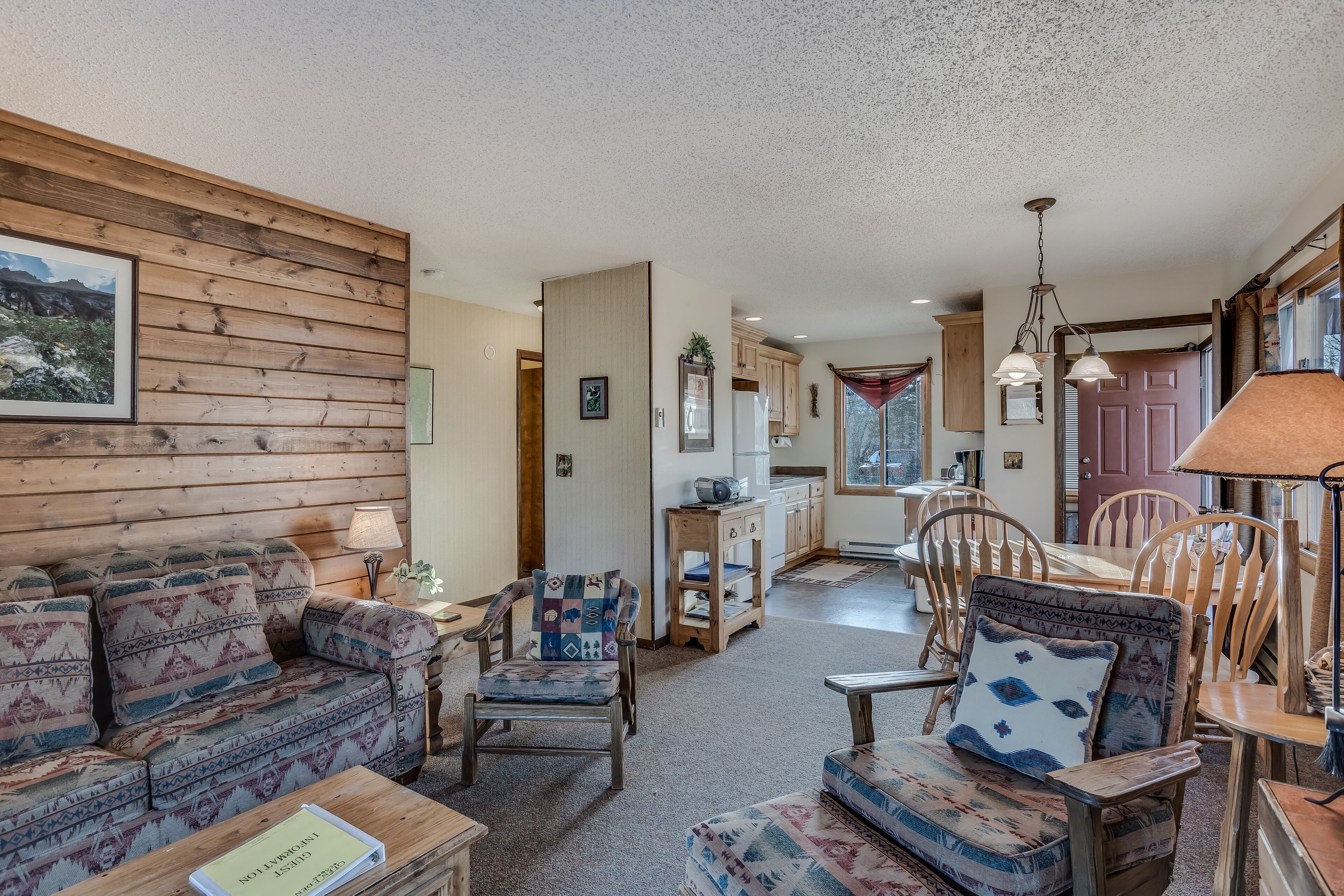 A cozy living room with a sofa, chairs, and a coffee table. There is a kitchen in the background with wooden cabinets and a dining table.