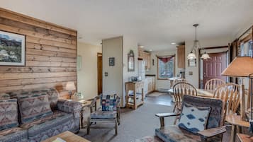 A cozy living room with a sofa, chairs, and a coffee table. There is a kitchen in the background with wooden cabinets and a dining table.