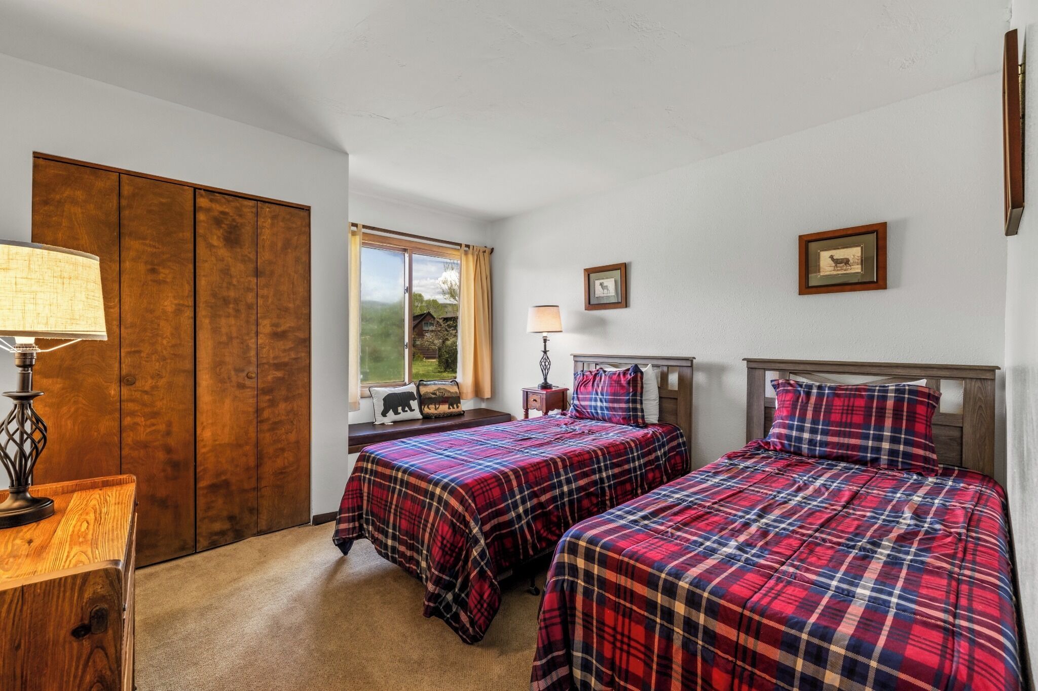 A bedroom with two beds, a wooden wardrobe, a window with curtains, and a lamp.