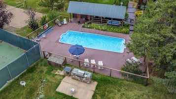 Una piscina rectangular con plataforma de madera, rodeada por una valla y una zona de barbacoa cubierta.