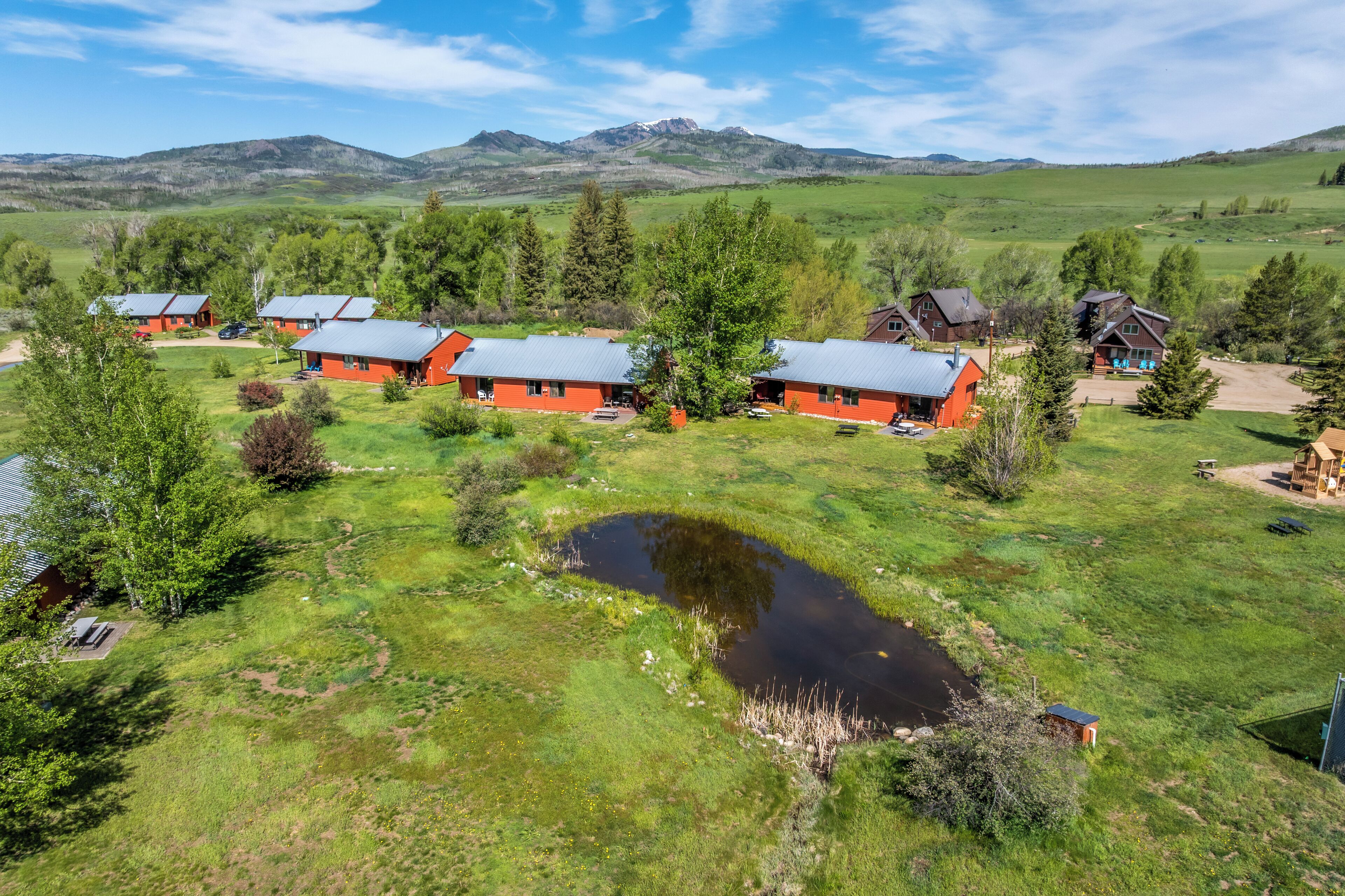 A cluster of red cabins with metal roofs, surrounded by green fields and a small pond, set against a backdrop of mountains.