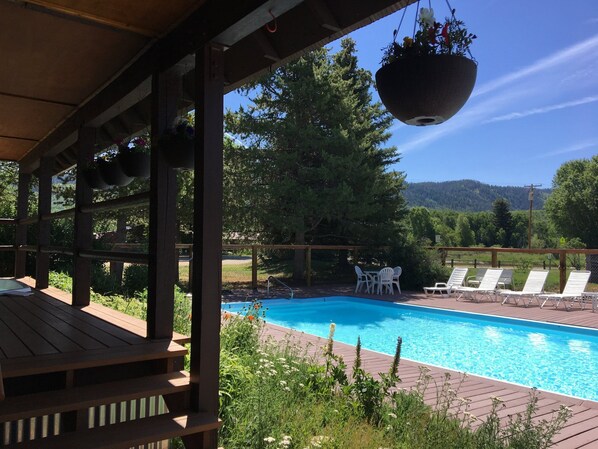 A wooden deck with a pool, white lounge chairs, and hanging flower baskets. - Homesteader - Peaceful cabin with beautiful views (Clark)