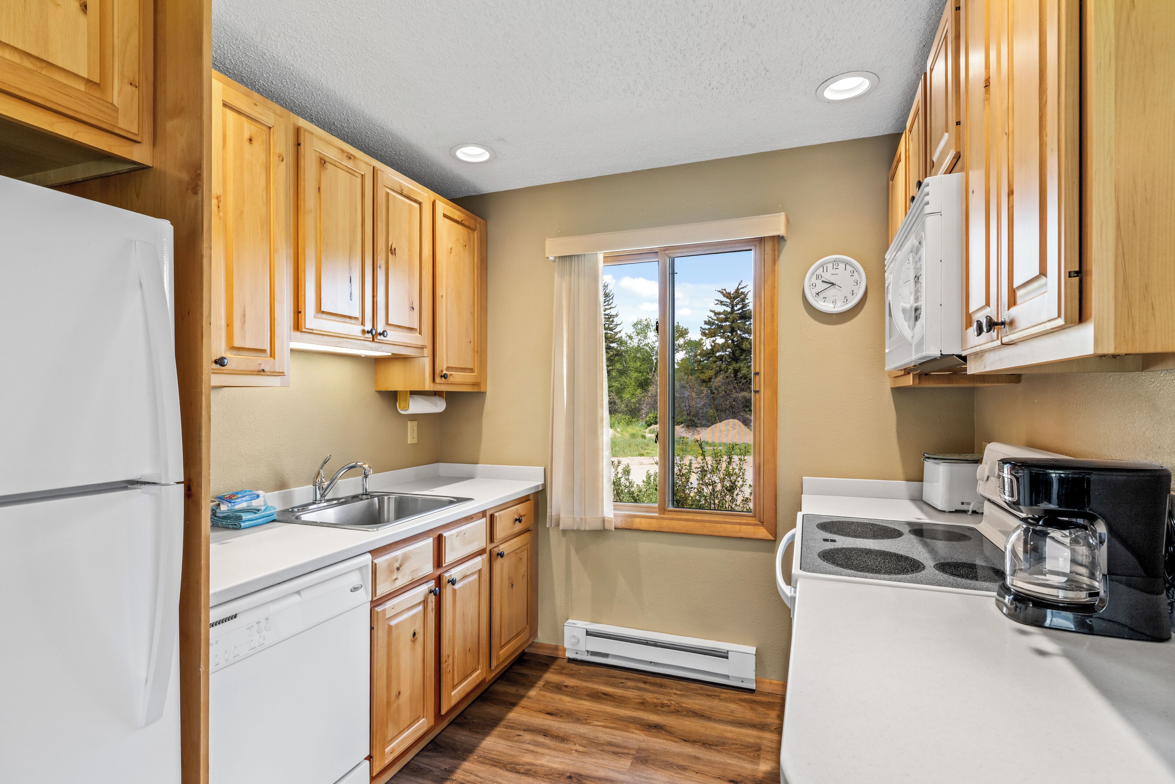 A kitchen with wooden cabinets, a white refrigerator, a stove, a sink, and a window with a view of trees.