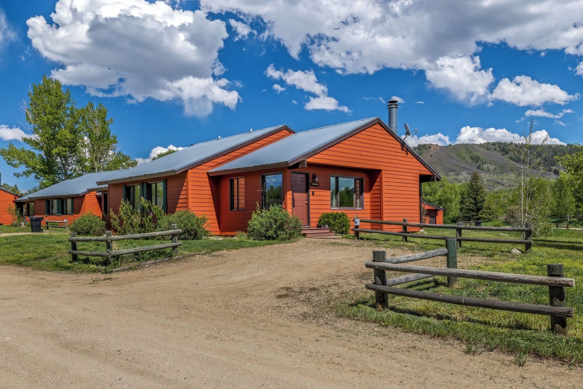Une cabane en bois rouge, surmontée d’un toit en métal, entourée d’une clôture en bois, située dans une zone herbeuse, avec des arbres et des montagnes en arrière-plan.