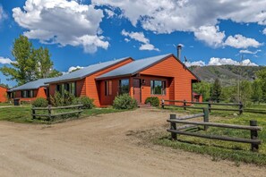 A red wooden cabin with a metal roof, surrounded by a wooden fence, set in a grassy area with trees and mountains in the background.