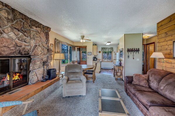 A cozy living room with a stone fireplace, a comfortable sofa, a dining area, and a kitchen in the background. - Hideaway - Cozy cabin in the great outdoors (Clark)