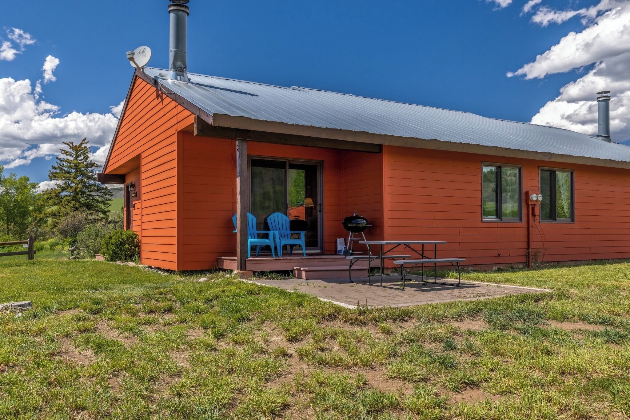 A single-story building with a red exterior, a covered patio area with a barbecue grill, and a blue chair.