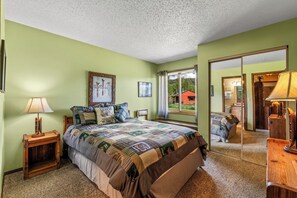 A bedroom with a bed, a nightstand, a lamp, and a window with a view of a red building. - Hideaway - Cozy cabin in the great outdoors (Clark)
