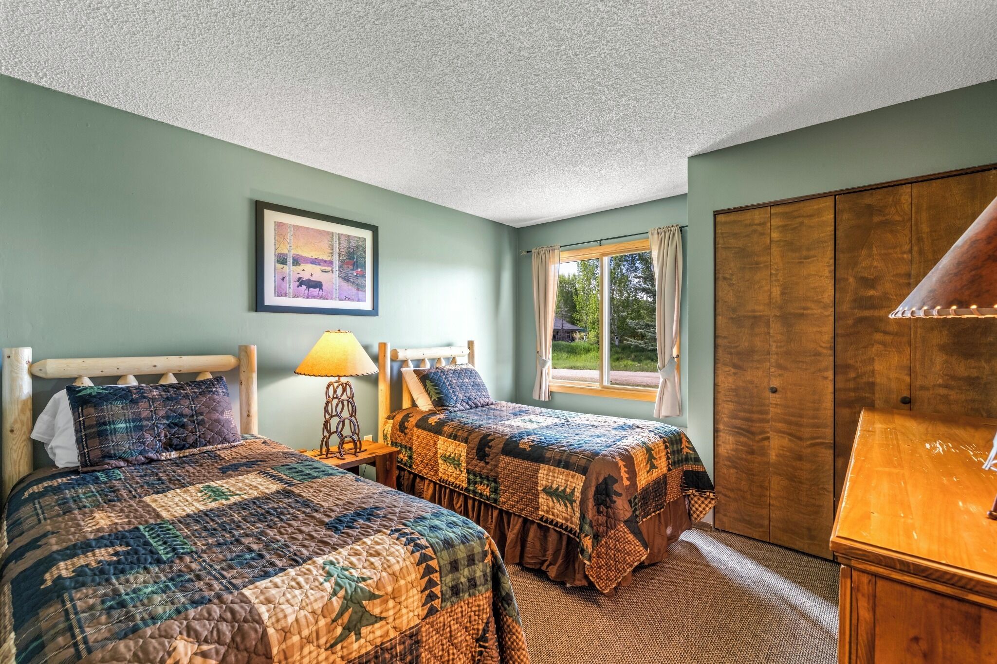 Une chambre avec deux lits, une tête de lit en bois, un tableau encadré, une fenêtre avec des rideaux et une armoire en bois.