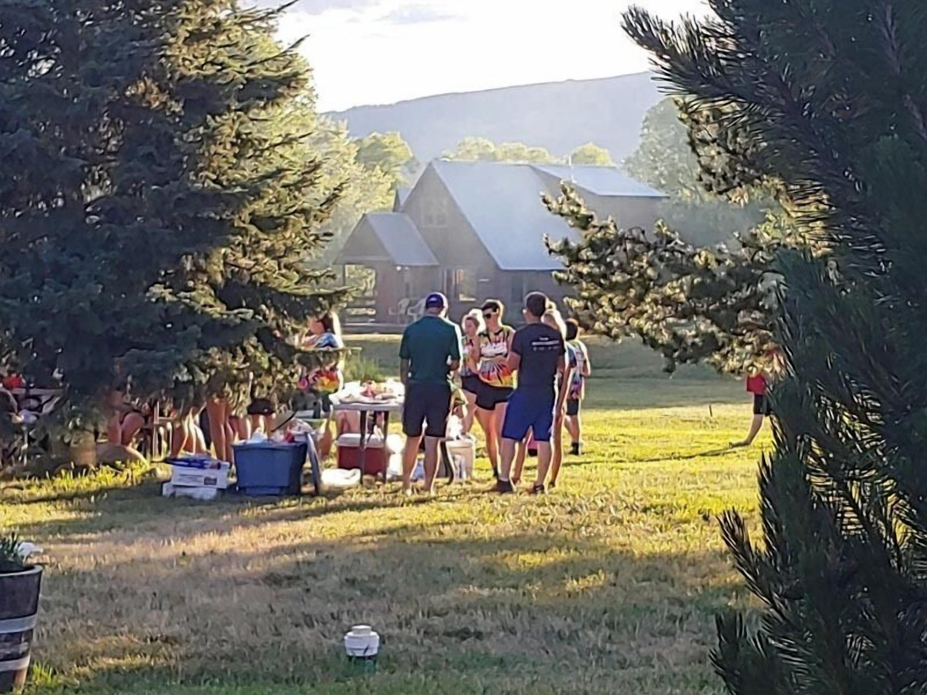 A group of people gathered around a table outdoors with a grassy field and trees in the background.