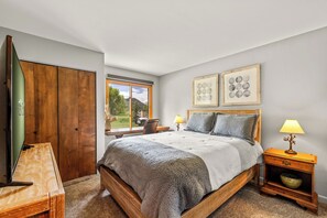 A bedroom with a wooden bed, two bedside tables with lamps, a window with a view, and two framed pictures on the wall. - Mustang - Escape to the wilderness (Clark)