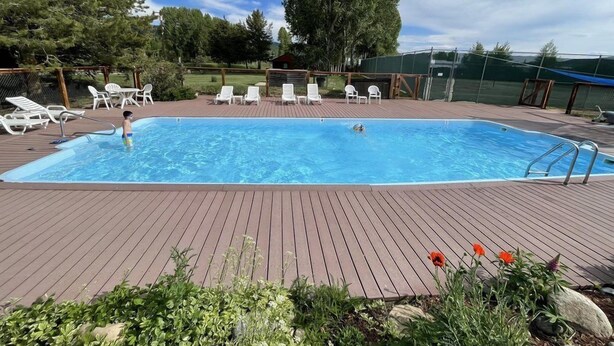A rectangular swimming pool with a wooden deck, surrounded by white lounge chairs, and a grassy area with trees and flowers. - Mustang - Escape to the wilderness (Clark)