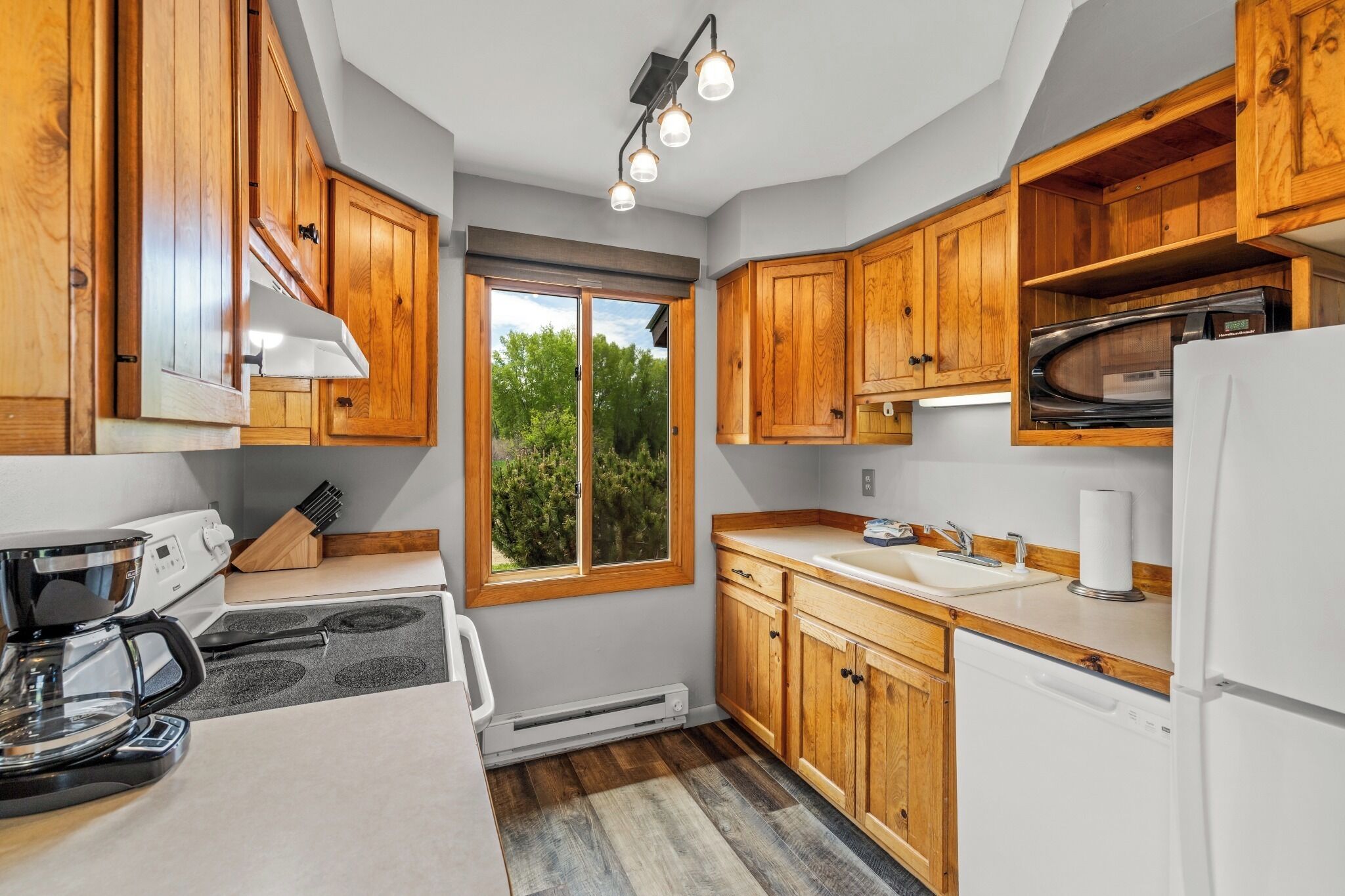 A compact kitchen with wooden cabinets, a built-in microwave, a coffee maker, and a window providing a view of greenery outside.