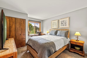 A bedroom with a wooden bed, two bedside tables with lamps, a window with a view, and two framed pictures on the wall. - Mustang - Escape to the wilderness (Clark)