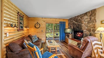 Interior acogedor de una cabaña de troncos con chimenea de piedra, muebles de madera y vistas al exterior.