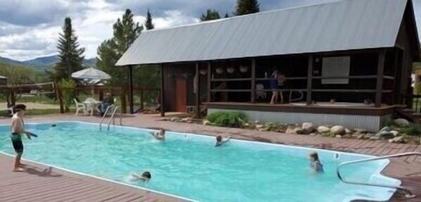 A swimming pool area with a covered deck, a building with large windows, and a mountainous backdrop.