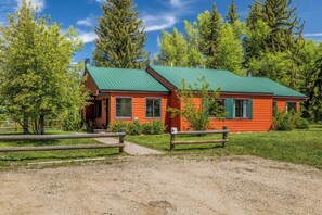 A single-story wooden cabin with a green roof, surrounded by a gravel driveway and a wooden fence. - Riverhouse - Fireplace, kitchen, pet friendly! (Clark)