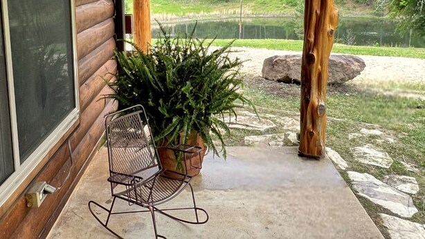 A wooden porch with a rocking chair and potted plants, overlooking a lake and forest. - Littlecreek: Rustic cabin get away. (Avinger)