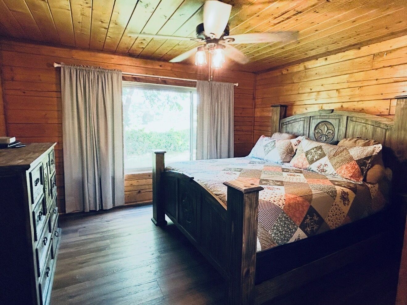 A wooden cabin bedroom with a bed, a dresser, a window with curtains, and a ceiling fan.