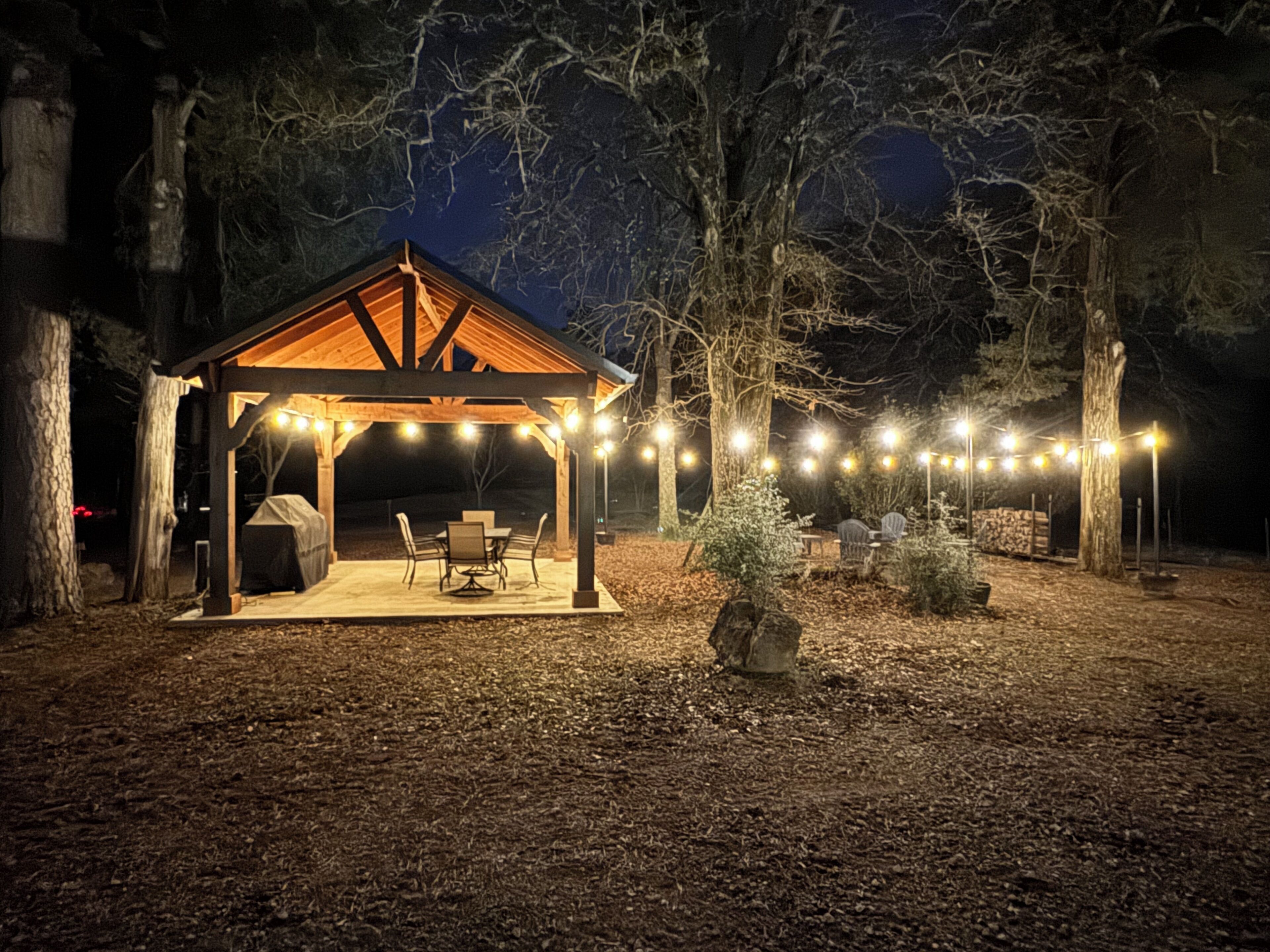 A covered outdoor seating area with string lights, surrounded by trees.