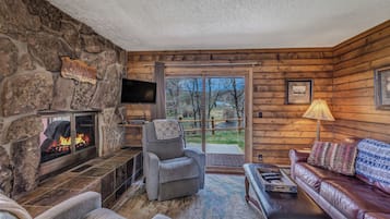 A cozy cabin interior with a stone fireplace, a comfortable seating area, and a large window offering a view of the outdoors.