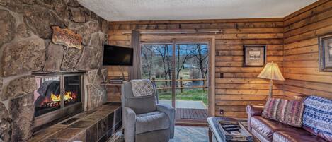 A cozy cabin interior with a stone fireplace, a comfortable seating area, and a large window offering a view of the outdoors.