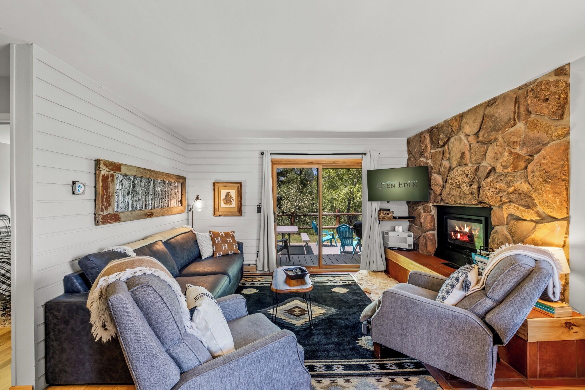 A living room with a stone wall, a fireplace, a TV, and a view of the outdoors.