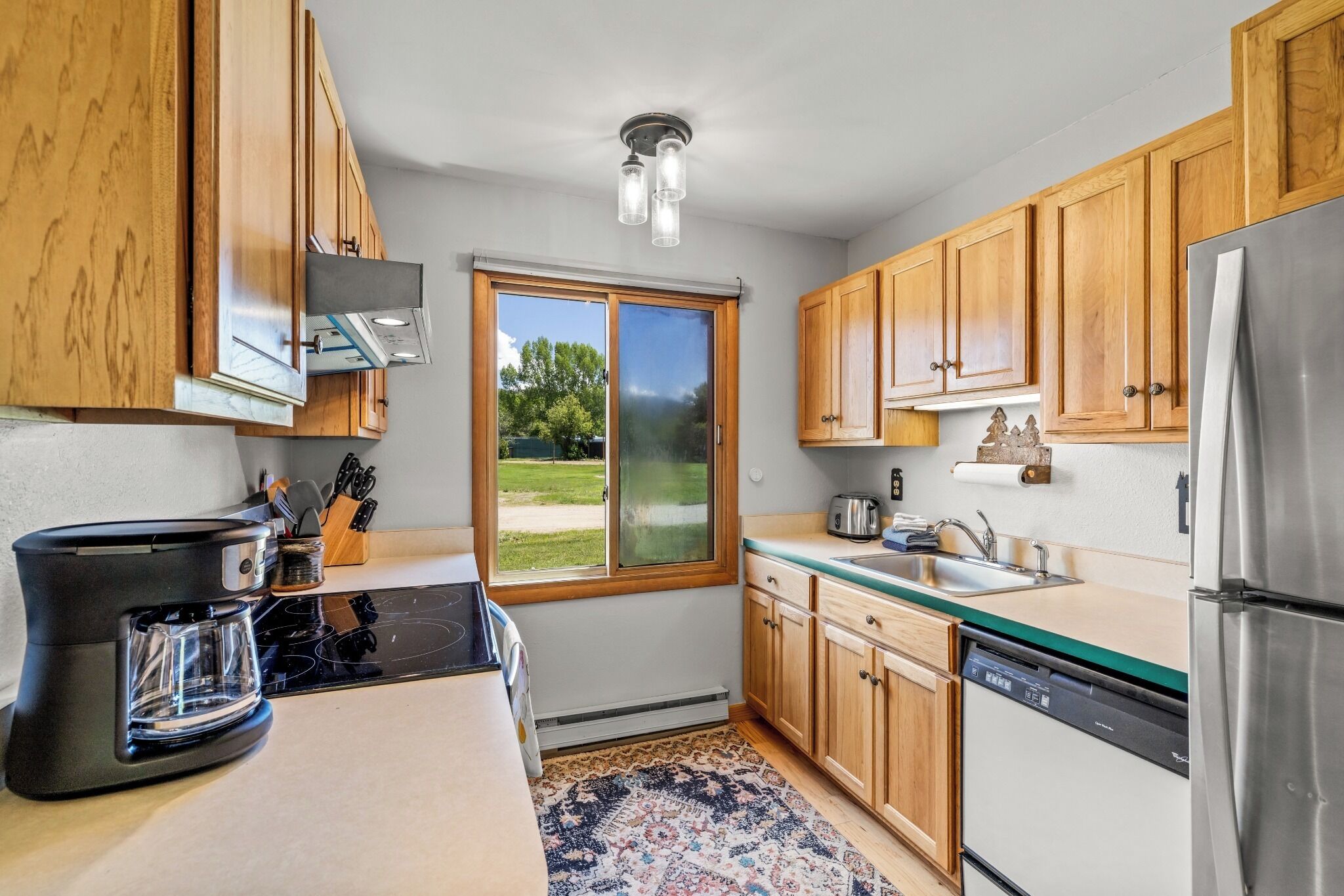 A kitchen with wooden cabinets, a stainless steel refrigerator, a coffee maker, and a window with a view of greenery.