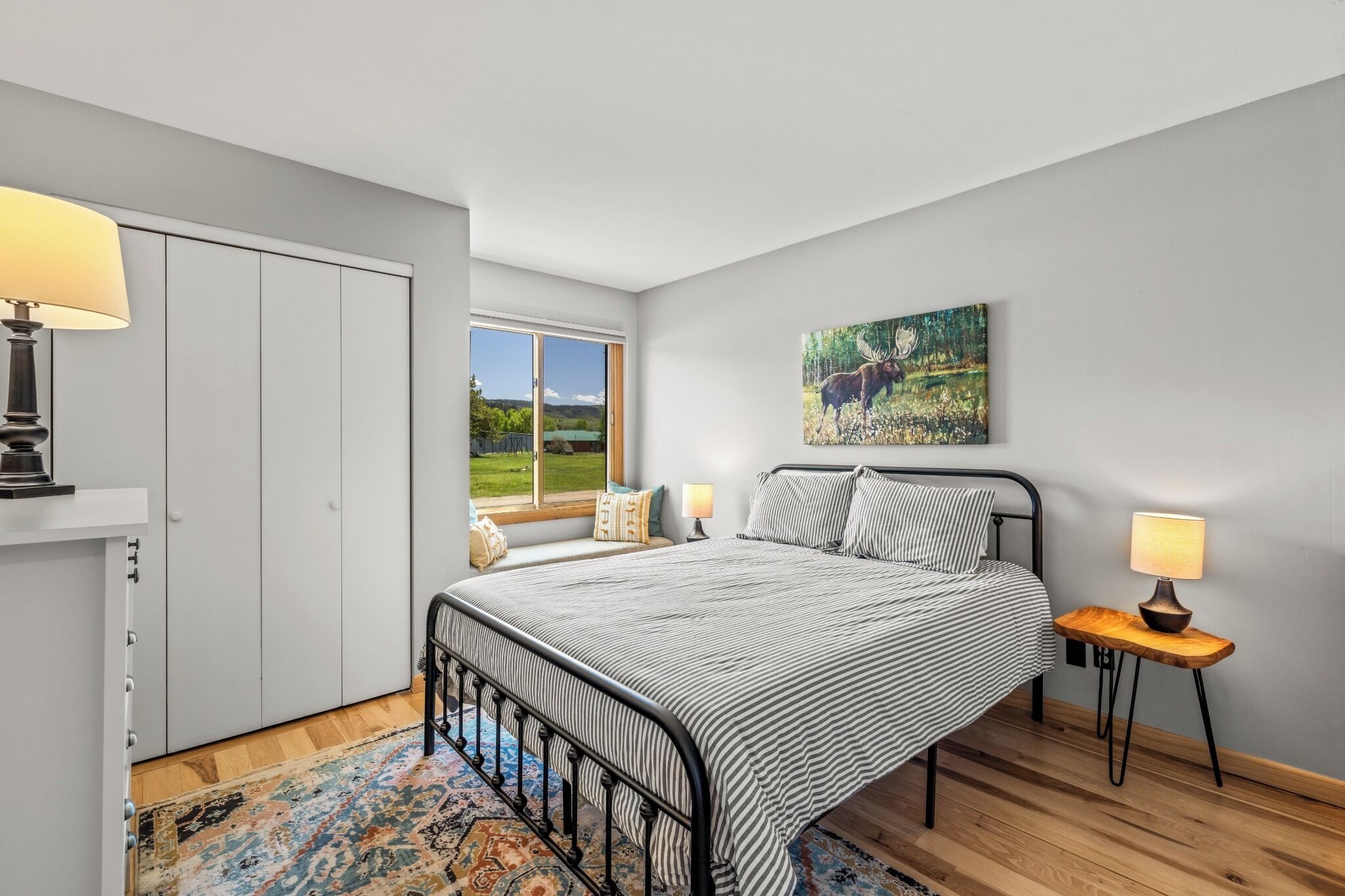 A bedroom with a bed, bedside lamps, a window with a view, a painting on the wall, and a wardrobe.